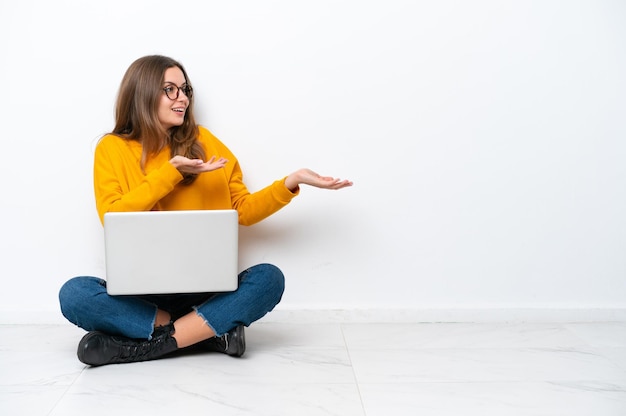 Giovane donna caucasica con un computer portatile seduto sul pavimento isolato su sfondo bianco con espressione facciale a sorpresa