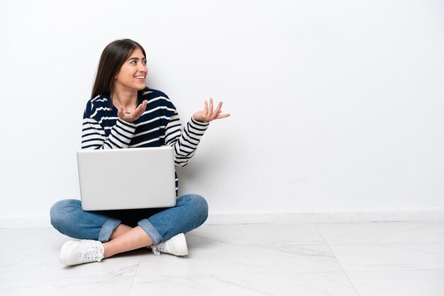 Giovane donna caucasica con un computer portatile seduto sul pavimento isolato su sfondo bianco con espressione facciale a sorpresa