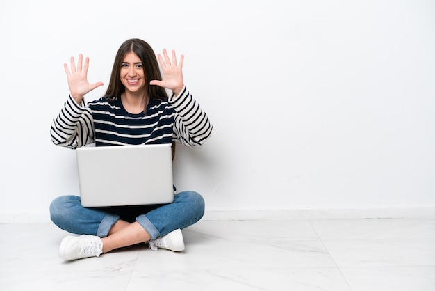 Giovane donna caucasica con un computer portatile seduto sul pavimento isolato su sfondo bianco contando dieci con le dita