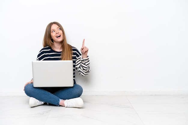 Giovane donna caucasica con un computer portatile seduto sul pavimento isolato su sfondo bianco rivolto verso l'alto e sorpreso