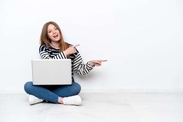 Giovane donna caucasica con un computer portatile seduto sul pavimento isolato su sfondo bianco sorpreso e indicando il lato