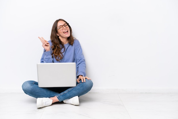 Giovane donna caucasica con un computer portatile seduto sul pavimento isolato su sfondo bianco sorridente e mostrando il segno di vittoria