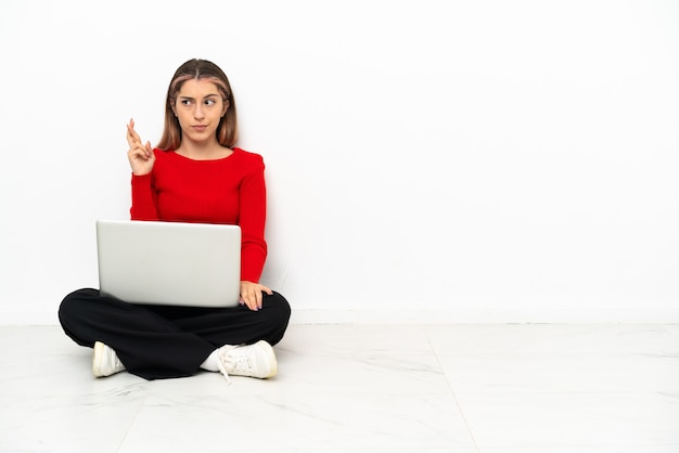 Giovane donna caucasica con un laptop seduto sul pavimento con le dita incrociate e che desiderano il meglio