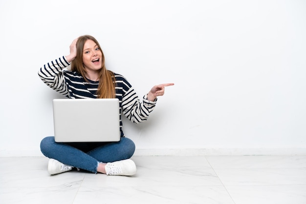 Giovane donna caucasica con un laptop seduto sul pavimento isolato su sfondo bianco sorpreso e puntando il dito di lato