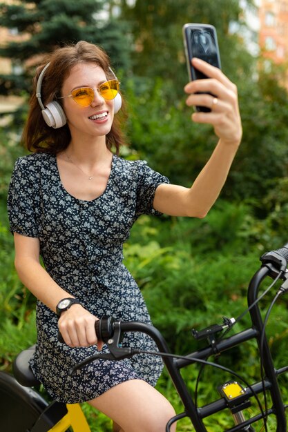 Giovane donna caucasica con un sorriso perfetto, labbra carnose, occhiali, auricolari, passeggiate nella natura, andare in bicicletta e registrare un video, fare selfie