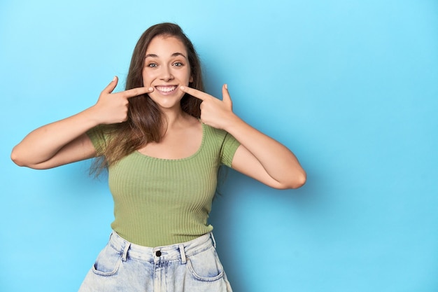 Giovane donna caucasica con un top verde su uno sfondo blu sorride indicando le dita alla bocca