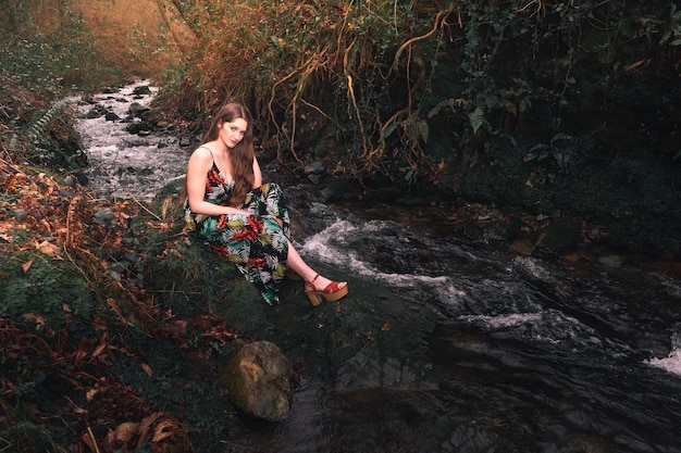 Giovane donna caucasica con un vestito colorato in mezzo alla foresta vicino a un piccolo fiume