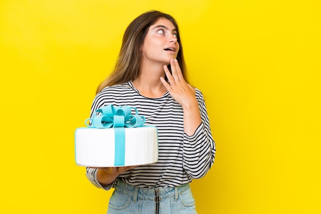 Giovane donna caucasica con una torta di compleanno isolata su uno sfondo giallo che alza lo sguardo mentre sorride