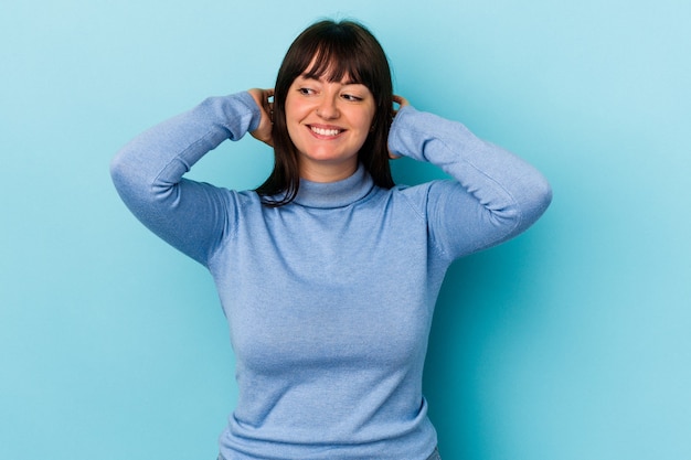 Giovane donna caucasica curvy isolata su fondo blu che si sente sicura, con le mani dietro la testa.