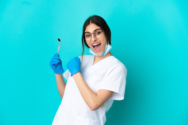 Giovane donna caucasica del dentista che tiene gli strumenti isolati su fondo blu che celebra una vittoria