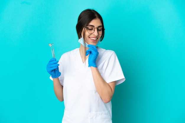 Giovane donna caucasica del dentista che tiene gli strumenti isolati su fondo blu che guarda al lato e che sorride