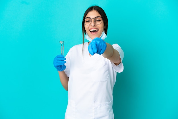 Giovane donna caucasica del dentista che tiene gli strumenti isolati su fondo blu che indica la parte anteriore con l'espressione felice