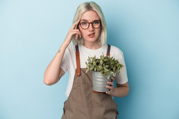 Giovane donna caucasica del giardiniere che tiene una pianta isolata su fondo blu che mostra un gesto di delusione con l'indice.