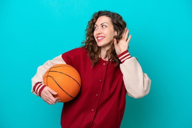 Giovane donna caucasica del giocatore di basket isolata su sfondo blu che ascolta qualcosa mettendo la mano sull'orecchio