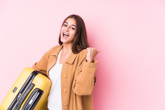 Giovane donna caucasica del viaggiatore che tiene i isolatedpoints di una valigia con il dito del pollice via, ridendo e spensierato.