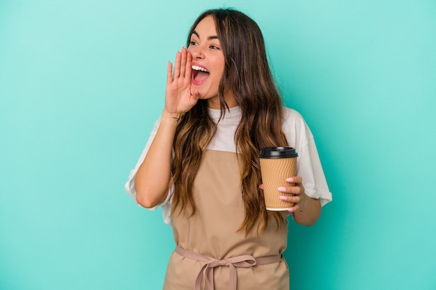 Giovane donna caucasica dell'addetto al negozio che tiene un caffè da asporto isolato su sfondo blu gridando e tenendo il palmo vicino alla bocca aperta.