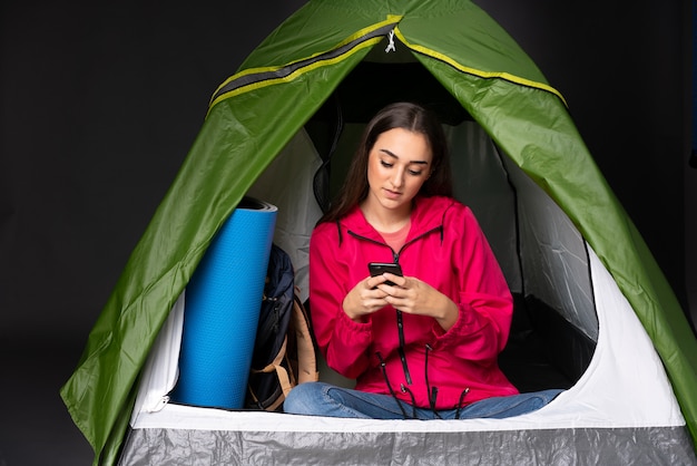 Giovane donna caucasica dentro una tenda verde di campeggio che invia un messaggio con il cellulare