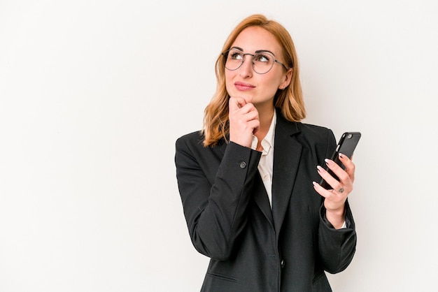 Giovane donna caucasica di affari che tiene il telefono cellulare isolato su sfondo bianco guardando di traverso con espressione dubbiosa e scettica.