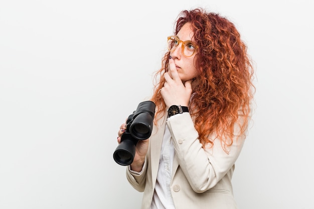 Giovane donna caucasica di redhead di affari che tiene un binocolo che guarda lateralmente con l'espressione dubbiosa e scettica.