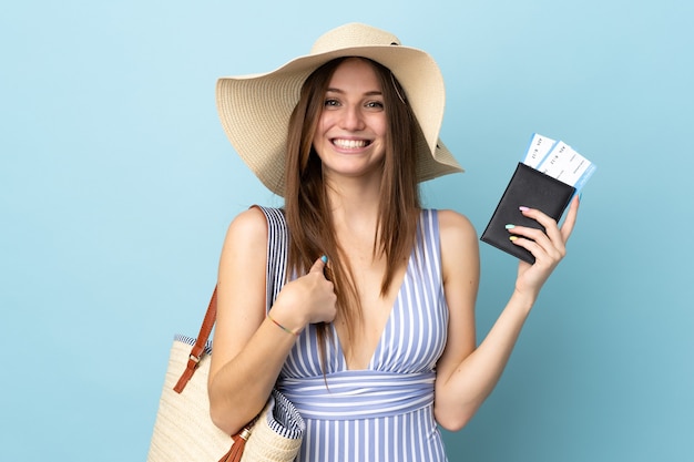 Giovane donna caucasica durante le vacanze estive in possesso di passaporto isolato su sfondo blu con espressione facciale a sorpresa