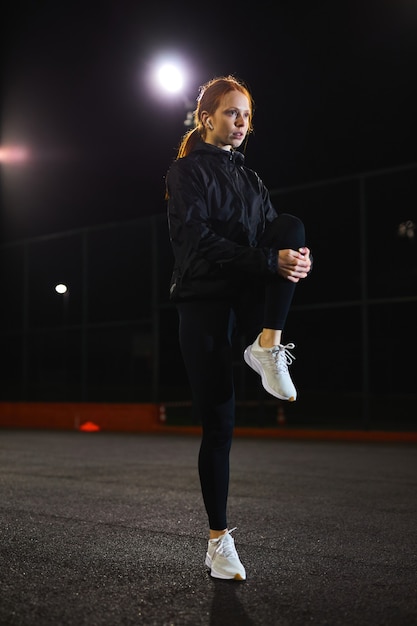 Giovane donna caucasica esile che fa allenamento di stretching all'aperto in una strada di notte di notte attraente...