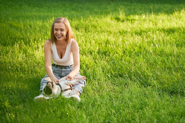 Giovane donna caucasica felice che si siede sull'erba con il bulldog francese maschio sulle gambe nel parco cittadino. Vista frontale della splendida ragazza che ride godendo del tramonto estivo con animali, accarezzando la sua pancia.