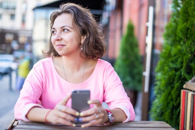 Giovane donna caucasica felice vaga che tiene smartphone in mani nel caffè della via di estate.