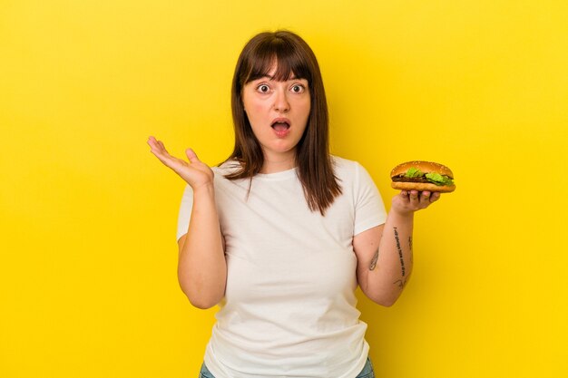 Giovane donna caucasica formosa che tiene hamburger isolato su sfondo giallo sorpreso e scioccato.