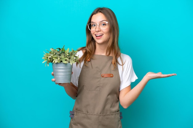 Giovane donna caucasica giardiniere che tiene una pianta isolata su sfondo blu con espressione facciale scioccata