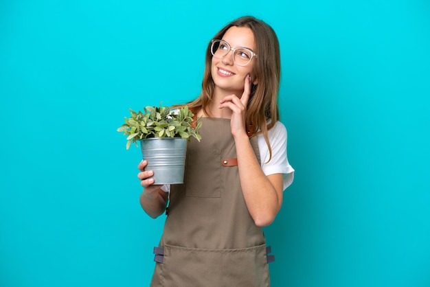 Giovane donna caucasica giardiniere che tiene una pianta isolata su sfondo blu pensando a un'idea mentre alza lo sguardo