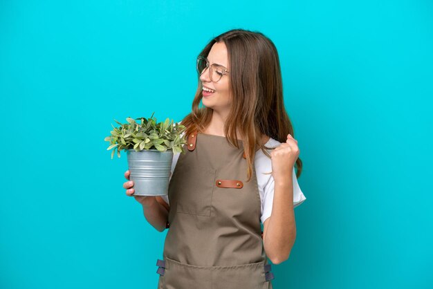 Giovane donna caucasica giardiniere in possesso di una pianta isolata su sfondo blu che celebra una vittoria