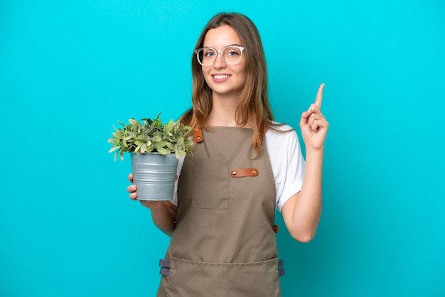 Giovane donna caucasica giardiniere in possesso di una pianta isolata su sfondo blu che indica una grande idea