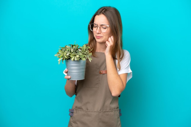 Giovane donna caucasica giardiniere in possesso di una pianta isolata su sfondo blu frustrata e che copre le orecchie