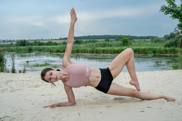 Giovane donna caucasica in abiti sportivi che fa yoga, si rilassa, si esercita vicino al lago, mattina per allenarsi in natura.