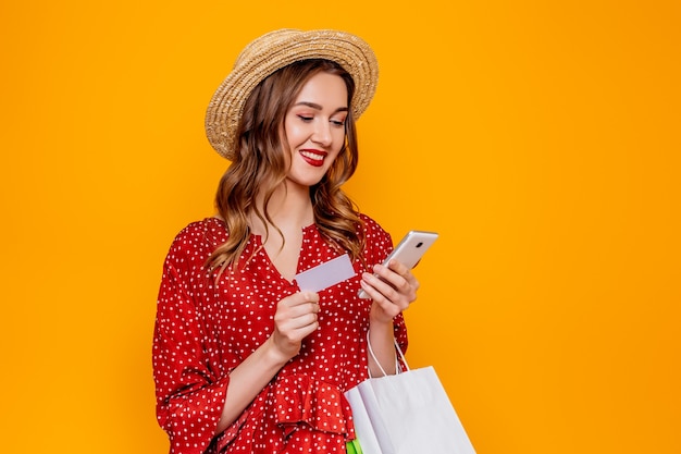 Giovane donna caucasica in abito estivo rosso con cappello di paglia sorridente e in possesso di carta di credito, telefono cellulare e pacchetto di acquisto ed esamina lo schermo del telefono isolato sul muro arancione dello studio