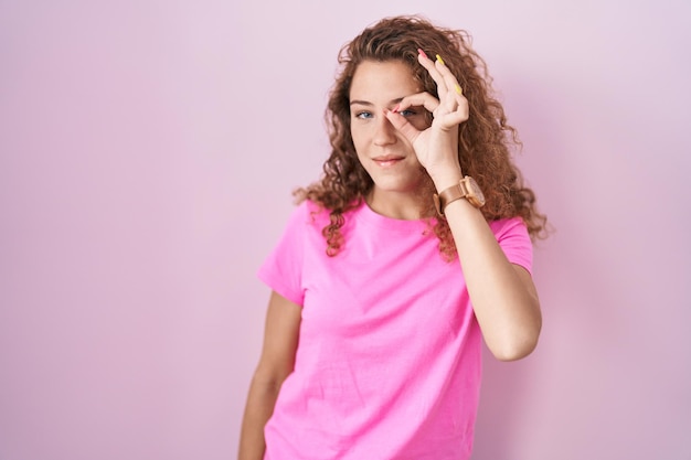 Giovane donna caucasica in piedi su sfondo rosa che fa gesto ok con la mano sorridente, occhio guardando attraverso le dita con faccia felice.