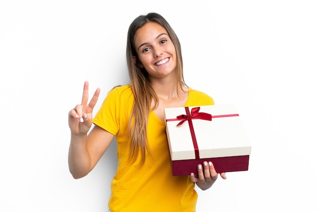 Giovane donna caucasica in possesso di un regalo isolato su sfondo bianco sorridente e mostrando il segno di vittoria