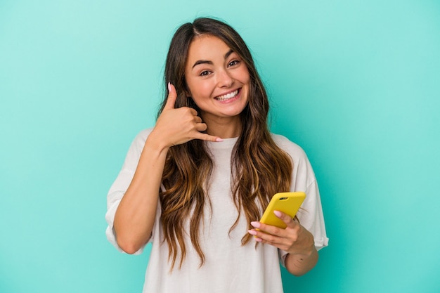 Giovane donna caucasica in possesso di un telefono cellulare isolato su sfondo blu che mostra un gesto di chiamata di telefonia mobile con le dita.