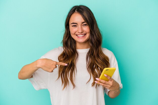 Giovane donna caucasica in possesso di un telefono cellulare isolato su sfondo blu persona che indica a mano uno spazio copia maglietta, orgogliosa e sicura di sé