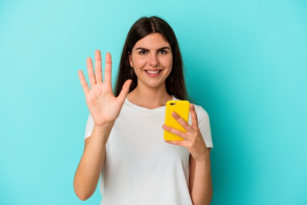 Giovane donna caucasica in possesso di un telefono cellulare isolato su sfondo blu sorridente allegro che mostra il numero cinque con le dita.
