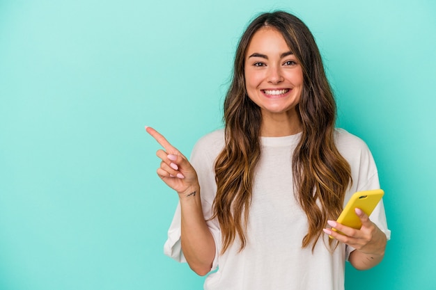 Giovane donna caucasica in possesso di un telefono cellulare isolato su sfondo blu sorridente e puntato da parte, mostrando qualcosa in uno spazio vuoto.