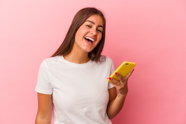 Giovane donna caucasica in possesso di un telefono cellulare isolato su sfondo rosa ridendo e divertendosi.