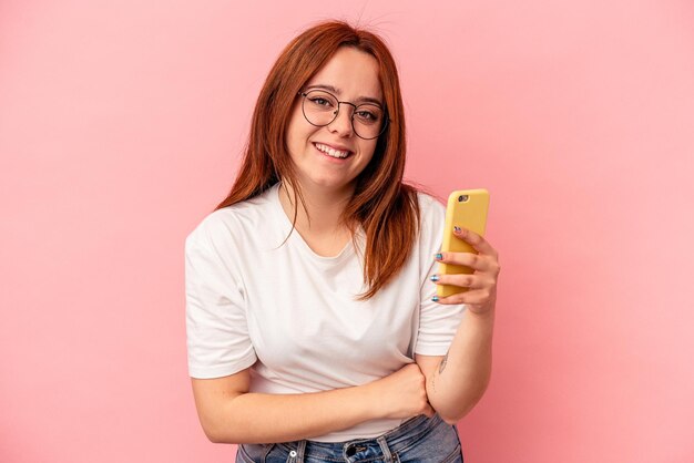 Giovane donna caucasica in possesso di un telefono cellulare isolato su sfondo rosa ridendo e divertendosi.