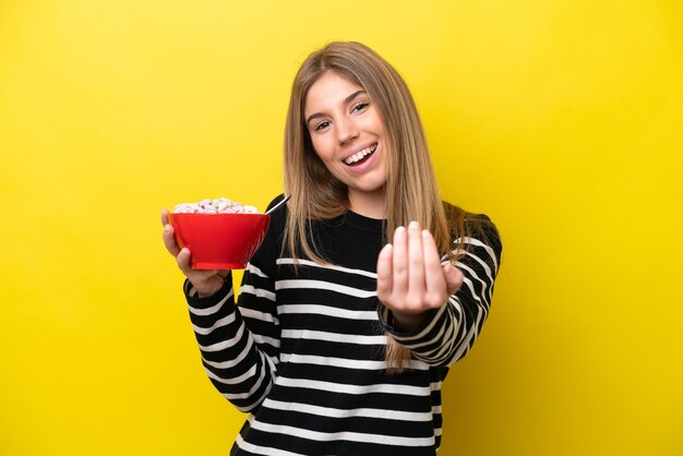 Giovane donna caucasica in possesso di una ciotola di cereali isolata su sfondo giallo che invita a venire con la mano Felice che tu sia venuto