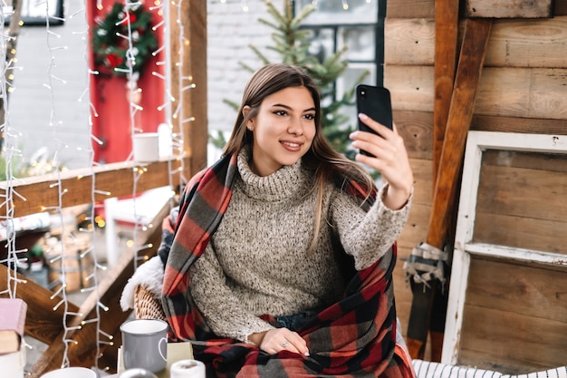 Giovane donna caucasica in un plaid che fa selfie sulla veranda con decorazioni natalizie.