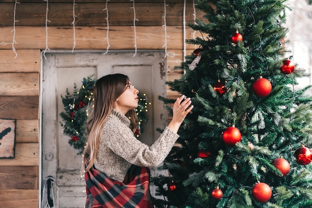 Giovane donna caucasica in un plaid decora un albero di Natale sulla veranda di una casa in legno.