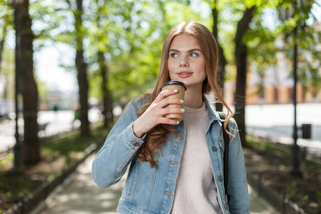 Giovane donna caucasica in una giacca di jeans beve caffè in viaggio in città