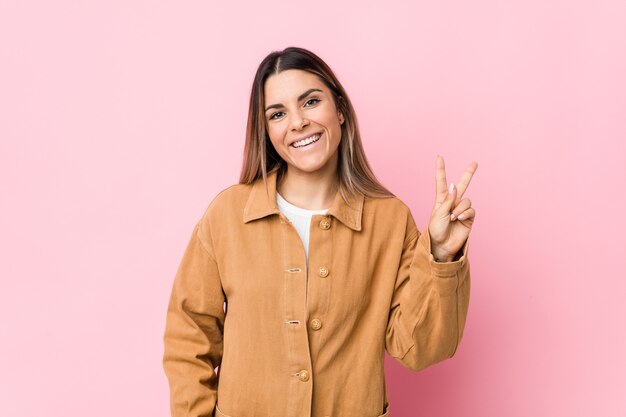 Giovane donna caucasica isolata che mostra il segno di vittoria e che sorride ampiamente.