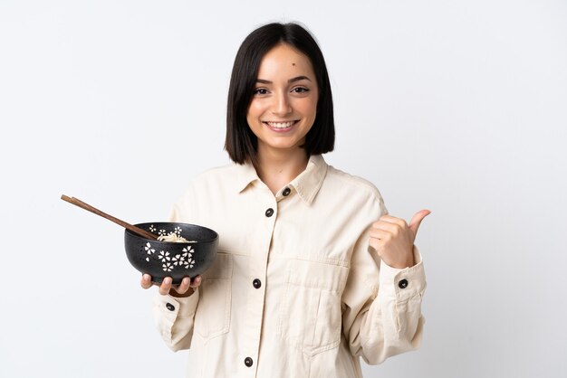 Giovane donna caucasica isolata su bianco che punta al lato per presentare un prodotto mentre si tiene una ciotola di spaghetti con le bacchette