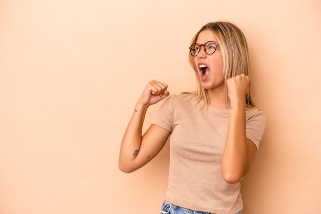 Giovane donna caucasica isolata su fondo beige che alza il pugno dopo una vittoria, concetto del vincitore.